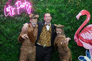 Three party goers standing in front of a green plant wall dressed up in jungle themed costumes.