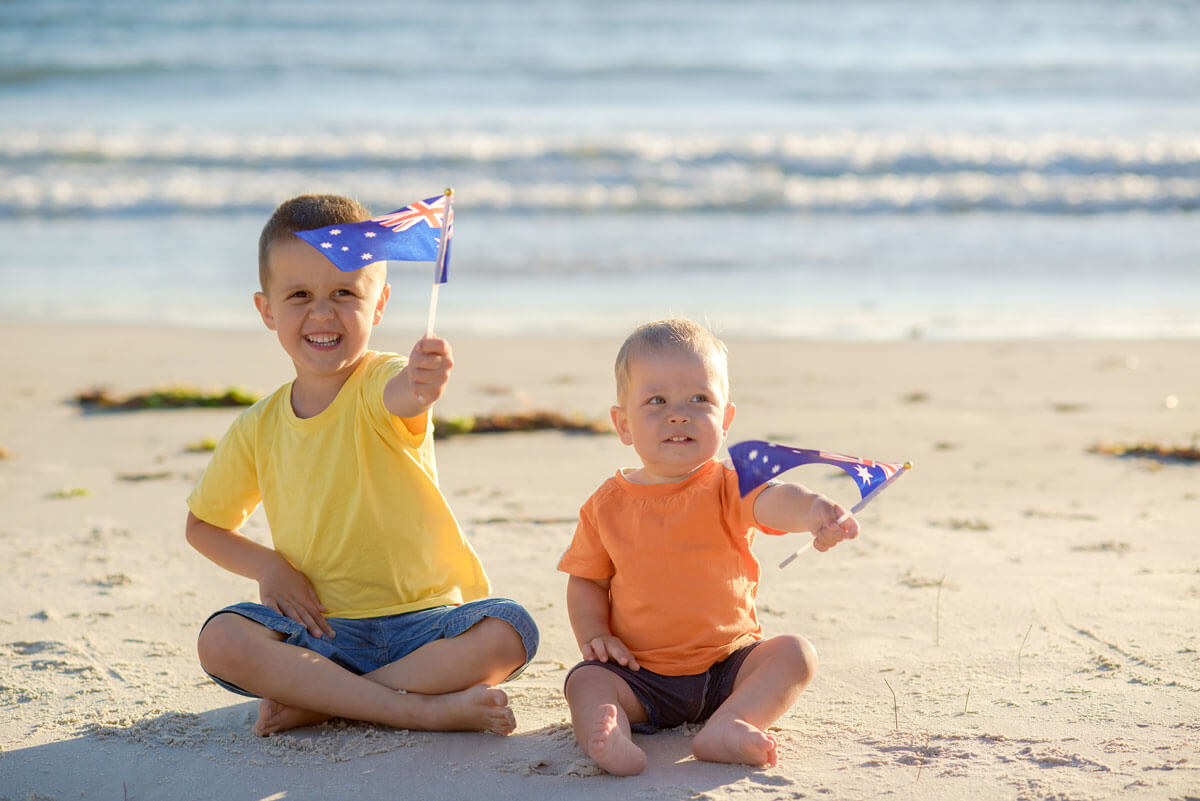 Two young toddler brothers sitting on the beach waving Australian flags
