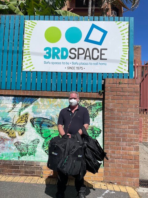 Man with mask standing infront of 3rdSpace sign holding a couple of One Light backpacks
